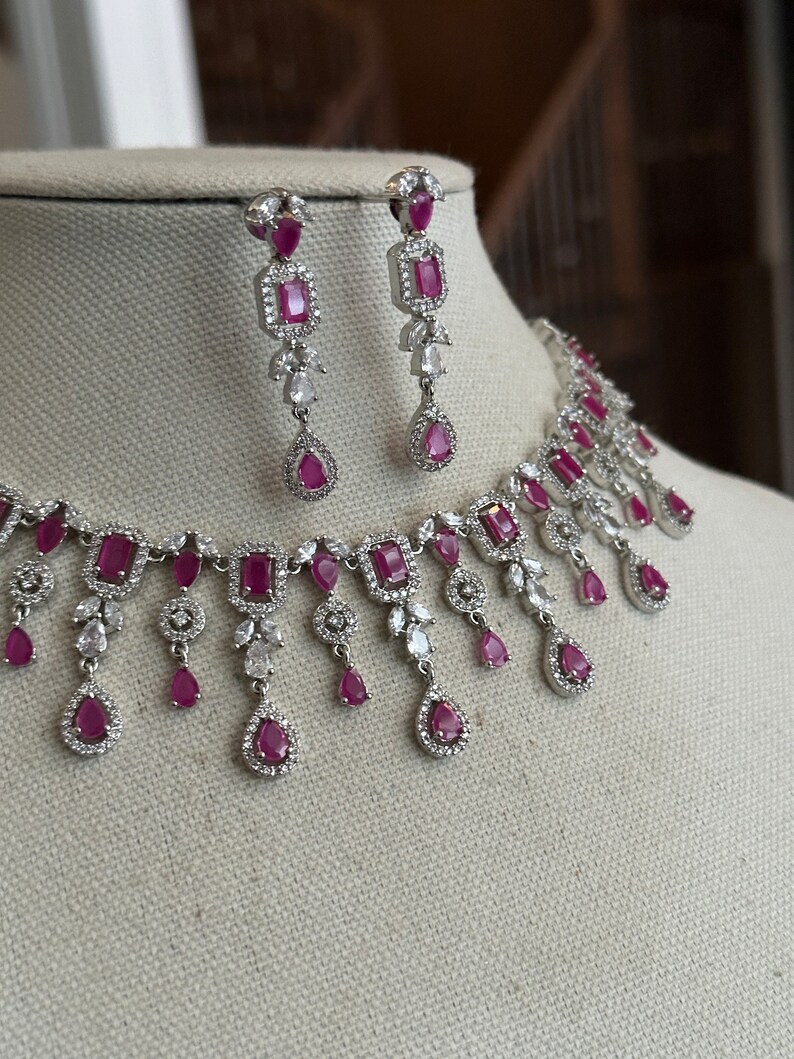 May include: A silver necklace with a row of pink and white stones, and matching earrings with pink stones. The necklace has a dangling design with multiple drops of pink and white stones.