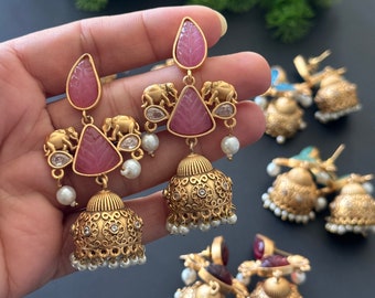 以下が含まれることがあります： ピンク色の宝石のアクセントと複雑なディテールが施されたゴールドトーンのイヤリング。 イヤリングは、しずく型のトップ、三角形のピンク色の石、小さな象のデザインが特徴です。 イヤリングは、小さな白い真珠のダングルが付いたベル型の底を持っています。
