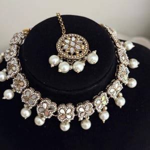 Puede incluir: Un conjunto de collar y tocado dorado. El collar presenta colgantes en forma de flor con centros espejados y perlas colgantes. El tocado tiene un colgante circular con un dise&ntilde;o similar, tambi&eacute;n con perlas.