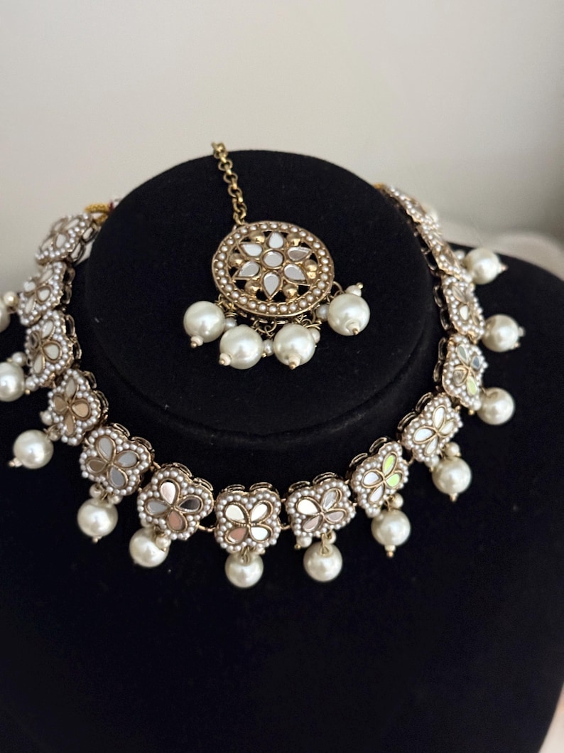 Puede incluir: Un conjunto de collar y tocado dorado. El collar presenta colgantes en forma de flor con centros espejados y perlas colgantes. El tocado tiene un colgante circular con un dise&ntilde;o similar, tambi&eacute;n con perlas.