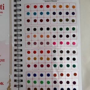 Op de afbeelding: Een spiraalgebonden boek met een verscheidenheid aan kleurrijke bindis. De bindis zijn gerangschikt in een rasterpatroon, met een spectrum aan kleuren, waaronder rood, blauw, groen en paars. De kaft van het boek toont de tekst "Hanskriti Bindies, Symbol of Beauty."