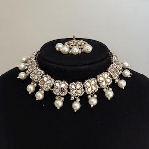 Puede incluir: Un collar plateado y un conjunto de tocado a juego. El collar presenta detalles florales espejados y perlas de imitaci&oacute;n colgantes. El tocado tiene un dise&ntilde;o similar con perlas de imitaci&oacute;n. El conjunto se exhibe sobre una cabeza de maniqu&iacute; negra.