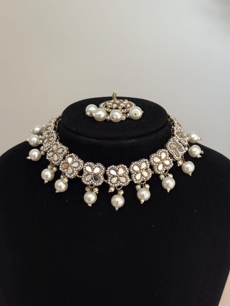 Puede incluir: Un collar plateado y un conjunto de tocado a juego. El collar presenta detalles florales espejados y perlas de imitaci&oacute;n colgantes. El tocado tiene un dise&ntilde;o similar con perlas de imitaci&oacute;n. El conjunto se exhibe sobre una cabeza de maniqu&iacute; negra.