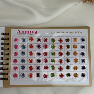 May include: A spiral-bound book featuring a variety of colorful Kumkum Bindi. The Ananya brand book displays rows of round, jewel-like bindis in shades of pink, blue, orange, purple, red, green, yellow, and navy blue. The text "EXCLUSIVE SPIRAL BOOK" is visible.