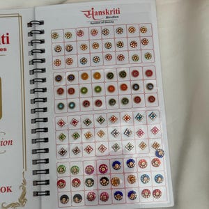 May include: A collection of colorful bindis, displayed in a spiral-bound book. The bindis feature various designs, including floral and geometric patterns, with a range of colors like red, blue, green, and gold. The brand name "Hanskriti Bindies" is visible.