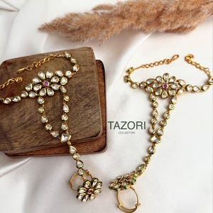 May include: Gold-toned hand jewelry with clear and red gemstones. The jewelry features a floral design with a ring attachment. The brand name "TAZORI COLLECTION" is visible in the image.