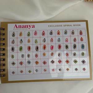 Puede incluir: Un libro con espiral titulado "Ananya KUMKUM BINDI" con una variedad de bindis coloridos y decorativos. Los bindis tienen forma de lágrima y cuadrada, con varios colores y detalles de joyas. El libro está sobre una superficie blanca. El texto "EXCLUSIVE SPIRAL BOOK" es visible.