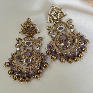 May include: A pair of gold-toned earrings with intricate designs and purple and white beads. The earrings feature a crescent moon shape with a floral pattern and a triangular top.