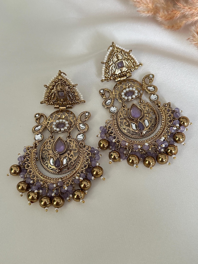 May include: A pair of gold-toned earrings with intricate designs and purple and white beads. The earrings feature a crescent moon shape with a floral pattern and a triangular top.