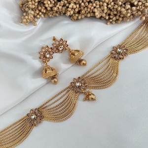 Könnte beinhalten: Ein goldfarbenes Schmuckset mit einer Halskette und passenden Ohrringen. Die Halskette hat mehrere Stränge und florale Akzente mit klaren Steinen. Die Ohrringe haben ein Blumenmuster mit einem glockenförmigen Anhänger. Das Set wird auf einer weißen Oberfläche präsentiert.