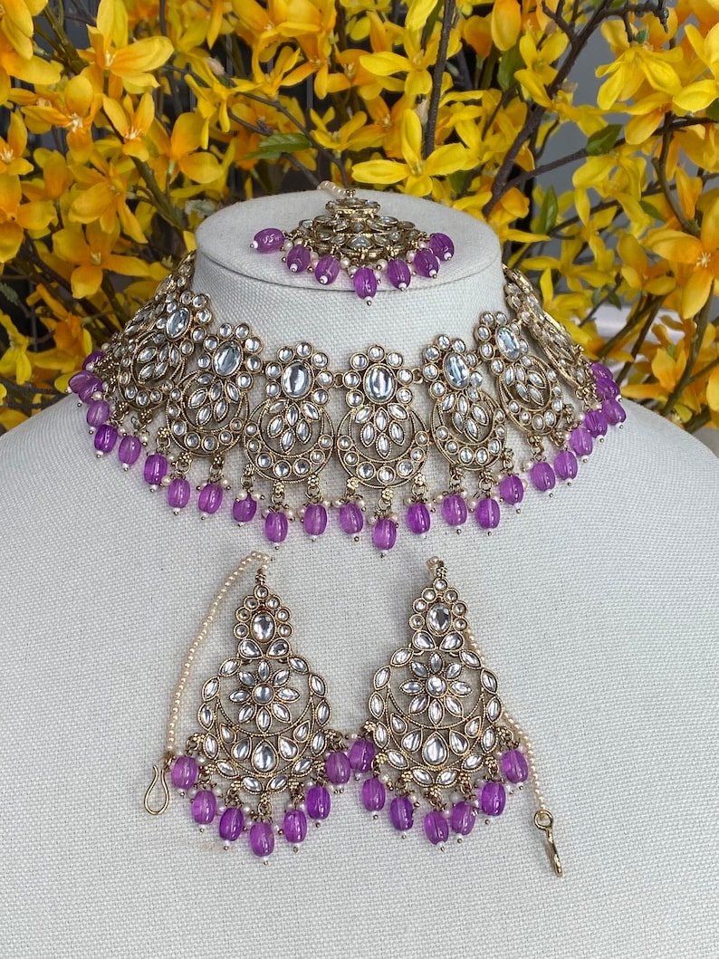 May include: A gold and silver necklace set with purple beads and clear stones. The necklace has a choker style and the earrings are dangle style.