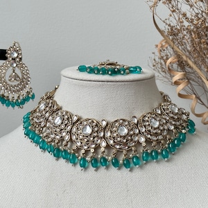 May include: A gold and silver necklace with a teardrop-shaped pendant and green beads. The necklace is on a white mannequin bust. The necklace is part of a jewelry set that includes earrings and a bracelet.