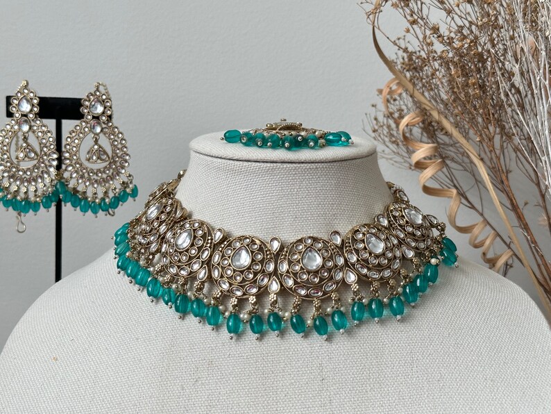 May include: A gold and silver necklace with a teardrop-shaped pendant and green beads. The necklace is on a white mannequin bust. The necklace is part of a jewelry set that includes earrings and a bracelet.