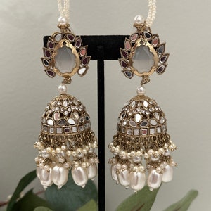 May include: A pair of gold tone earrings with a teardrop shaped clear stone, surrounded by smaller mirrored stones and pink and purple accents. The earrings have a dangling section with multiple pearl beads.