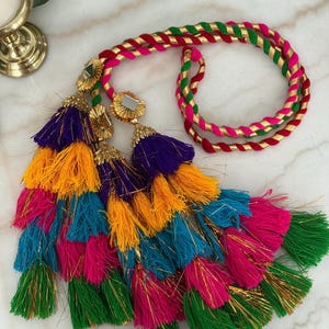 May include: A decorative accessory featuring a braided cord in pink, green, and gold, attached to a cluster of multi-colored tassels in purple, yellow, teal, pink, and green, accented with gold-toned metal caps and small square mirrors.