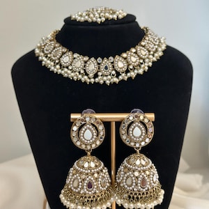 以下が含まれることがあります： ゴールドとホワイトのインドのブライダルジュエリーセット。チョーカーネックレス、イヤリング、マングティッカ。ネックレスとイヤリングは、鏡面仕上げの涙型の装飾とパールをあしらっています。