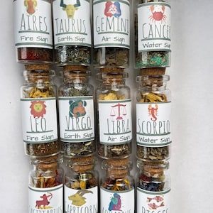 May include: Twelve small glass bottles with cork tops, each filled with different colored glitter and dried herbs. Each bottle has a label with a zodiac sign, name, and element. The signs are: Aries, Taurus, Gemini, Cancer, Leo, Virgo, Libra, Scorpio, Sagittarius, Capricorn, Aquarius, and Pisces.