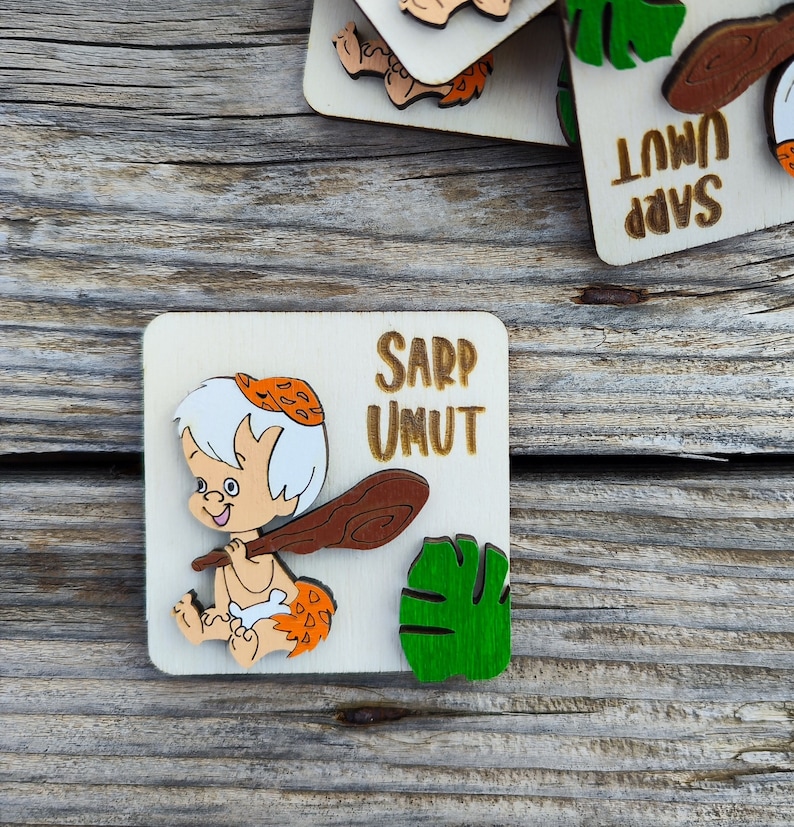 Puede incluir: Una placa de madera con una imagen de dibujos animados de un hombre de las cavernas sosteniendo un garrote. La placa est&aacute; decorada con una hoja verde y el texto "Sarp Umut".