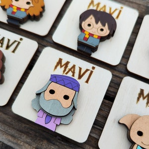 May include: Wooden square tags with the name "Mavi" written on them. Each tag features a different cartoon character from the Harry Potter series. The characters include Harry Potter, Hermione Granger, Ron Weasley, and Dumbledore.