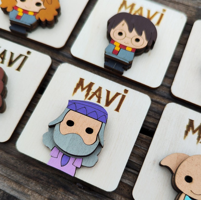 May include: Wooden square tags with the name "Mavi" written on them. Each tag features a different cartoon character from the Harry Potter series. The characters include Harry Potter, Hermione Granger, Ron Weasley, and Dumbledore.