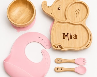 May include: A baby feeding set featuring a wooden bowl, an elephant-shaped plate, a bib, and utensils. The plate and utensils are engraved with the name "Mia". The set includes a pink bib and pink silicone suction cups on the bowl and plate.
