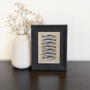 May include: A framed vertical print featuring six stacked blue and silver fish illustrations on a neutral background, housed in a black wooden frame. Beside the frame sits a matte white ceramic vase containing delicate dried baby's breath flowers.