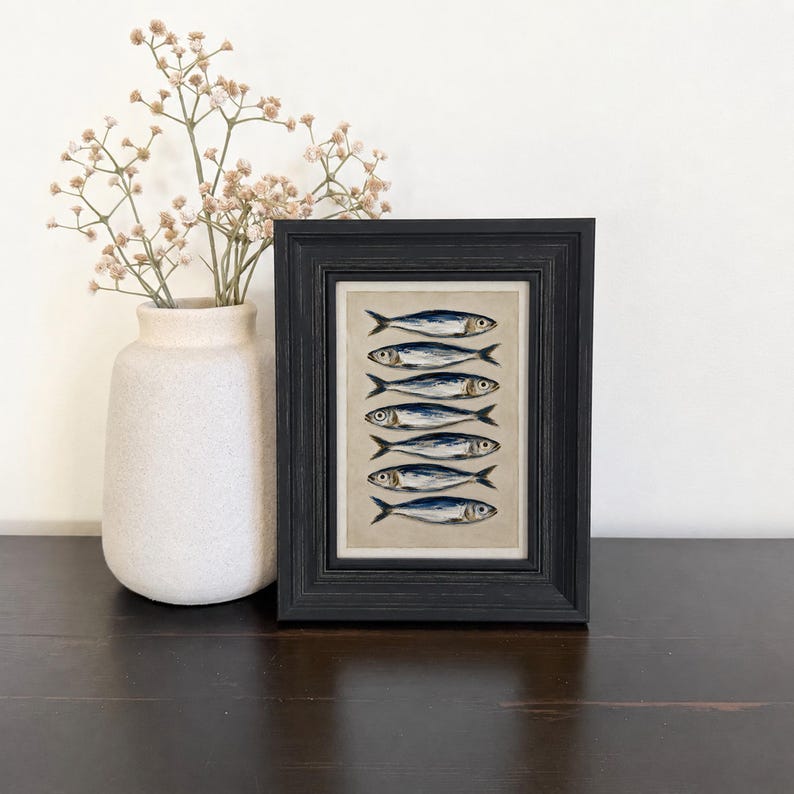 May include: A framed vertical print featuring six stacked blue and silver fish illustrations on a neutral background, housed in a black wooden frame. Beside the frame sits a matte white ceramic vase containing delicate dried baby's breath flowers.