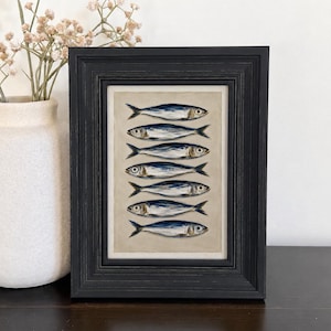 May include: A framed artwork featuring a vertical stack of seven painted fish in shades of blue, silver, and white against a neutral beige background. The piece is enclosed in a dark, textured wooden frame with a stepped profile and a white mat.