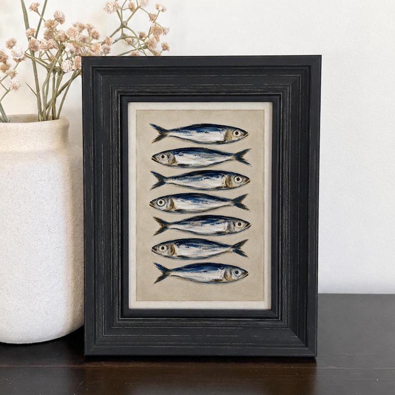 May include: A framed artwork featuring a vertical stack of seven painted fish in shades of blue, silver, and white against a neutral beige background. The piece is enclosed in a dark, textured wooden frame with a stepped profile and a white mat.