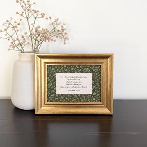 May include: A gold-toned rectangular picture frame containing a white paper insert with black text that reads: For I know the plans I have for you, declares the Lord, plans to prosper you and not to harm you, plans to give you hope and a future. Jeremiah 29:11.