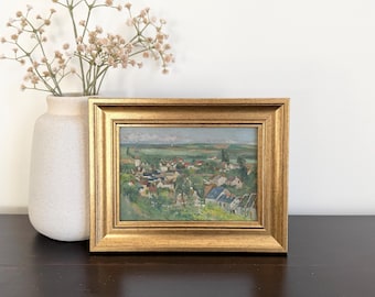 May include: A landscape painting in a rectangular gold frame depicting a small village with houses, trees, and green fields under a cloudy sky. The artwork features muted tones of green, blue, and brown, capturing a rural scene with a distant horizon.
