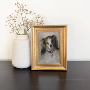 May include: A framed oil painting of a small dog with long, dark ears and a white face, wearing a collar with a small tag. The painting is encased in a rectangular, ornate gold-toned frame, positioned next to a matte white vase with dried flowers.