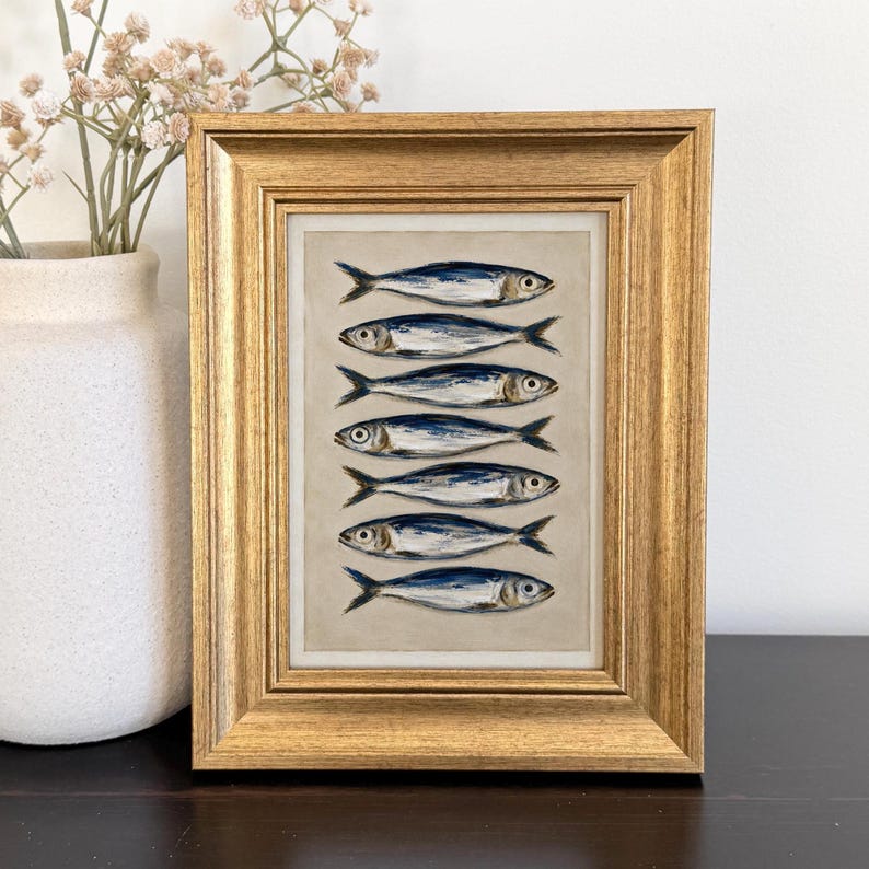May include: A framed artwork featuring a vertical stack of seven painted fish in shades of blue, silver, and white against a neutral beige background. The piece is enclosed in a textured, metallic gold frame with a classic, multi-layered border design.