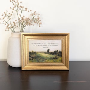 May include: A rectangular gold-toned frame containing a landscape print with the text "THIS IS THE DAY THE LORD HAS MADE; LET US REJOICE AND BE GLAD IN IT" and "Psalm 118:24" printed above a green field and trees under a pale sky.