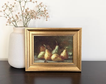 May include: An oil painting in a gold-toned frame depicting a still life of five pears in shades of yellow, green, and brown. The artwork features a dark, moody background and is displayed next to a light-colored vase containing dried flowers.