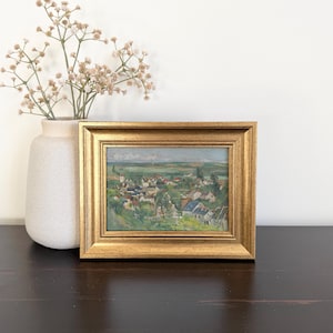 May include: A landscape painting in a thick, ornate gold-colored frame depicting a small village with houses, trees, and green fields under a soft sky. The artwork is displayed next to a light-colored ceramic vase containing dried beige flowers.