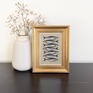 May include: A framed artwork featuring a vertical arrangement of seven blue and silver fish illustrations on a neutral background, encased in a textured gold frame. The artwork is displayed next to a light beige vase containing dried baby's breath.