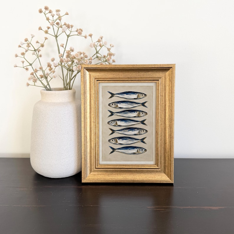 May include: A framed artwork featuring a vertical arrangement of seven blue and silver fish illustrations on a neutral background, encased in a textured gold frame. The artwork is displayed next to a light beige vase containing dried baby's breath.