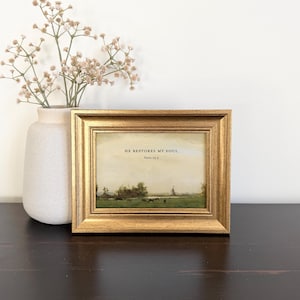 May include: A landscape art print in a gold-toned frame featuring a green field with trees and a windmill under a pale sky. The text HE RESTORES MY SOUL Psalm 23:3 is printed in black in the center. A light-colored vase with dried flowers sits nearby.