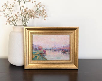 May include: A framed landscape painting featuring a river scene with soft pink, blue, and green brushstrokes, housed in a metallic gold frame. Beside the painting stands a matte off-white ceramic vase containing a sprig of dried baby's breath.