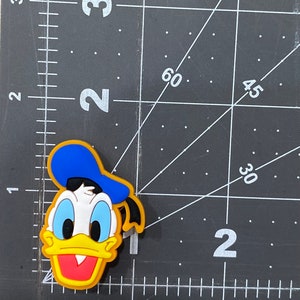 Może przedstawiać: Ż&oacute;łta, niebieska i czerwona postać z kresk&oacute;wki z niebieskim kapeluszem i ż&oacute;łtym dziobem. Postać jest gumową zawieszką.