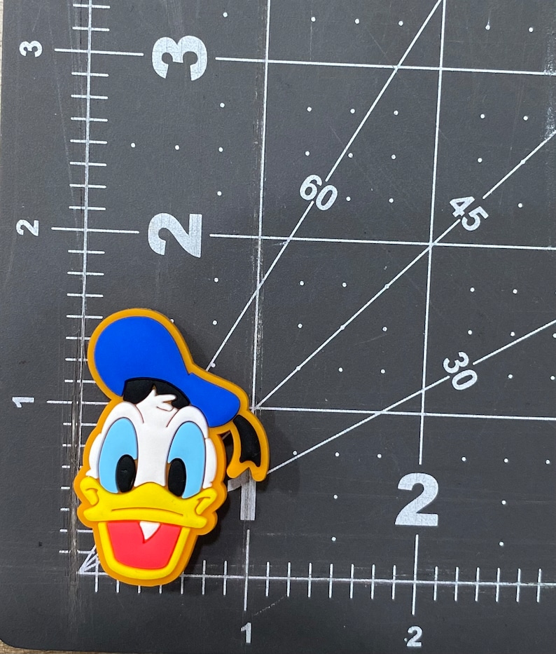 Może przedstawiać: Ż&oacute;łta, niebieska i czerwona postać z kresk&oacute;wki z niebieskim kapeluszem i ż&oacute;łtym dziobem. Postać jest gumową zawieszką.