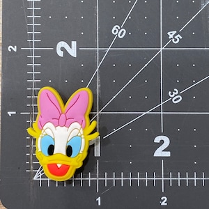 Może przedstawiać: Ż&oacute;łty gumowy brelok z postacią kresk&oacute;wkowej kaczki z r&oacute;żową kokardką, niebieskimi oczami i otwartym czerwonym dziobem. Kaczka ma na sobie biały kołnierzyk.