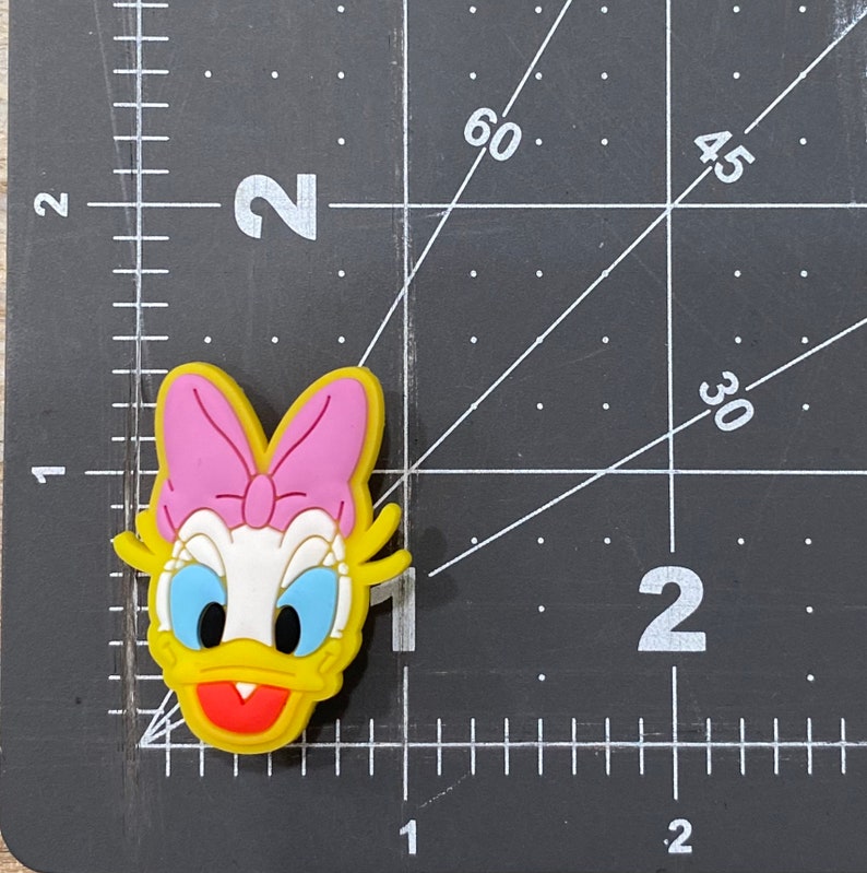 Może przedstawiać: Ż&oacute;łty gumowy brelok z postacią kresk&oacute;wkowej kaczki z r&oacute;żową kokardką, niebieskimi oczami i otwartym czerwonym dziobem. Kaczka ma na sobie biały kołnierzyk.