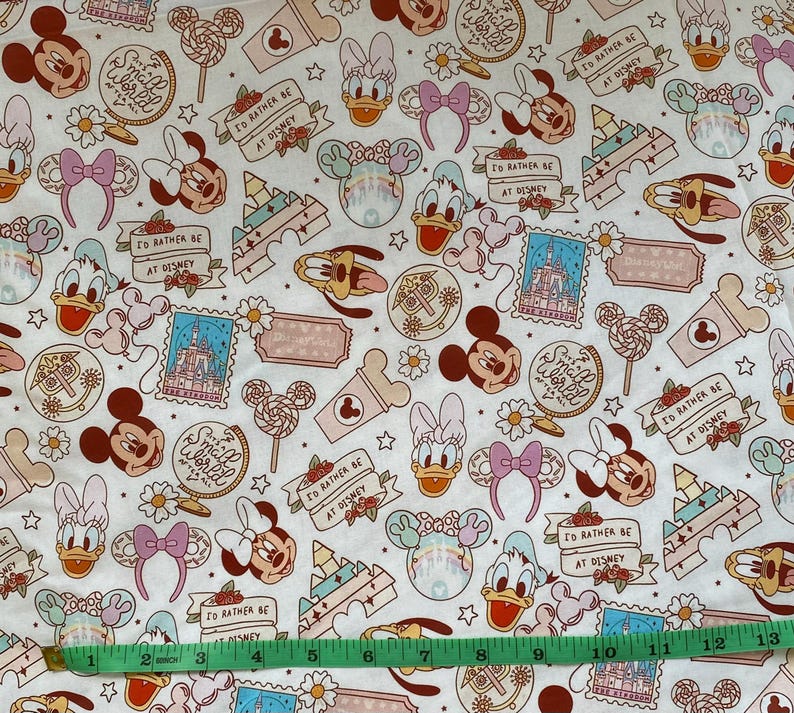 May include: White fabric with a repeating pattern of cartoon characters, including Mickey Mouse, Minnie Mouse, Donald Duck, and Goofy. The fabric features a variety of colors, including pink, red, blue, and yellow. The fabric also includes the text "Small World" and "I'd rather be at Disney".