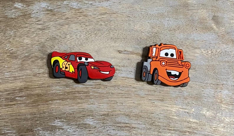 May include: Two red and orange cartoon car figurines. The red car is a race car with the number 95 on the side. The orange car is a tow truck with a smiling face.