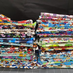 以下が含まれることがあります： 漫画のキャラクターが印刷されたカラフルな布の正方形の積み重ね。布は2つの積み重ねに配置されており、左側に1つ、右側に1つあります。布は、青、緑、赤、黄、紫など、さまざまな色を混ぜています。