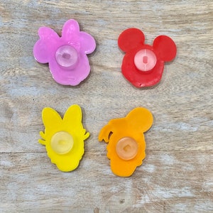 Może przedstawiać: Cztery kolorowe, plastikowe przyssawki w kształcie postaci. Postacie to Myszka Miki, Minnie Mouse, Donald Duck i Daisy Duck. Przyssawki są czerwone, r&oacute;żowe, ż&oacute;łte i pomarańczowe.