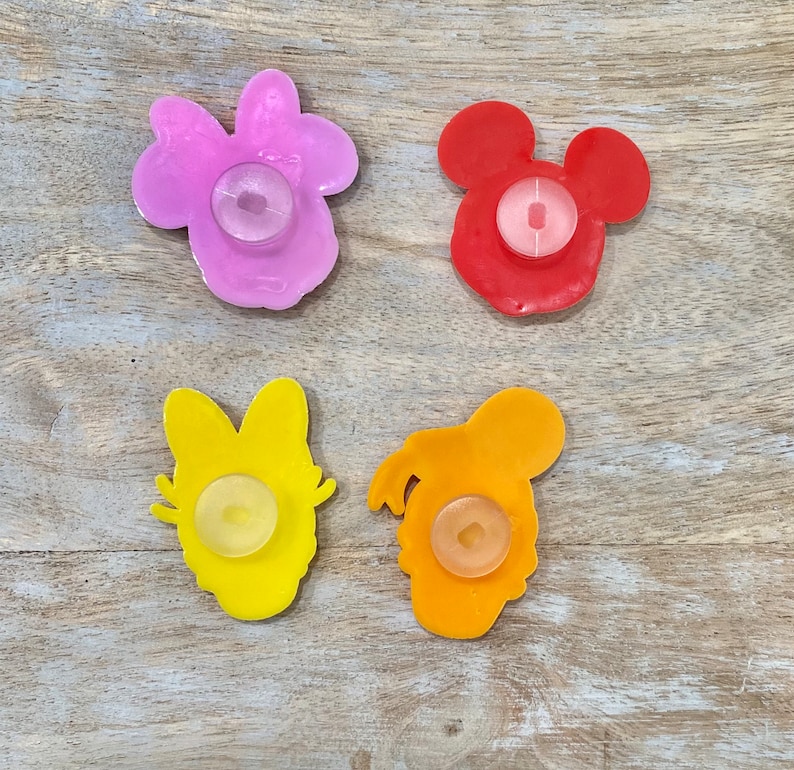 Może przedstawiać: Cztery kolorowe, plastikowe przyssawki w kształcie postaci. Postacie to Myszka Miki, Minnie Mouse, Donald Duck i Daisy Duck. Przyssawki są czerwone, r&oacute;żowe, ż&oacute;łte i pomarańczowe.