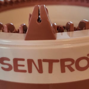 Op de afbeelding: Close-up van een witte plastic breimachine met een bruine plastic bovenkant en het woord "SENTRO" gedrukt op de voorkant.